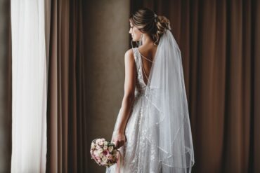 Bride in a lace gown and long veil holds a pink rose bouquet while standing beside a softly lit window.