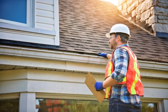 Roofing contractor in hard hat and safety vest inspects a home's shingle roof while holding a clipboard by the gutter.