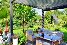 A backyard surrounded by greenery, with solar panels and an aluminum pergola over a table with multiple chairs.