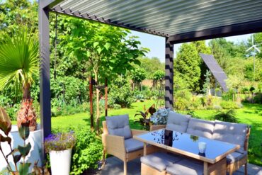 A backyard surrounded by greenery, with solar panels and an aluminum pergola over a table with multiple chairs.