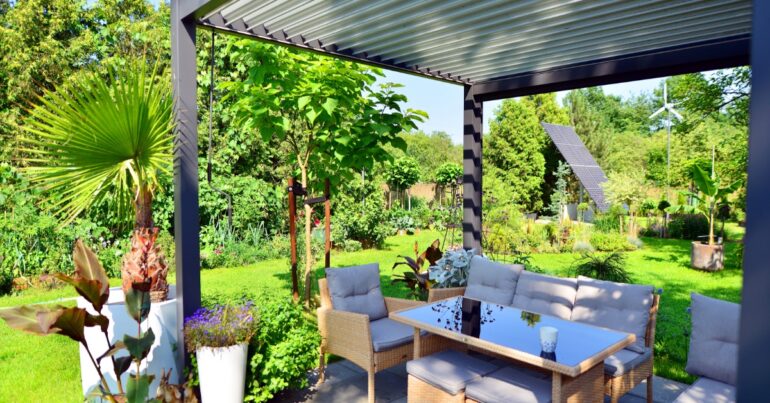 A backyard surrounded by greenery, with solar panels and an aluminum pergola over a table with multiple chairs.