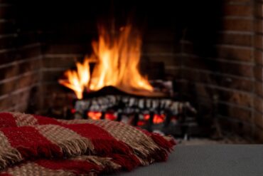 A brick fireplace with wood burning under a large flame indoors next to a red and brown blanket placed on a grey bench.
