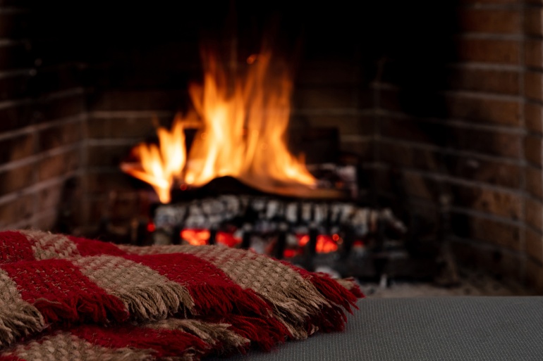 A brick fireplace with wood burning under a large flame indoors next to a red and brown blanket placed on a grey bench.