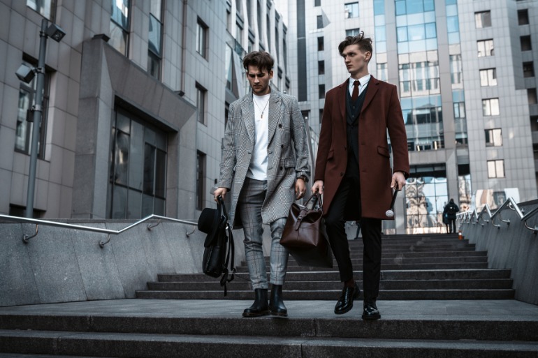 Two men wearing long coats while carrying leather bags, walking down concrete steps outdoors next to a building.