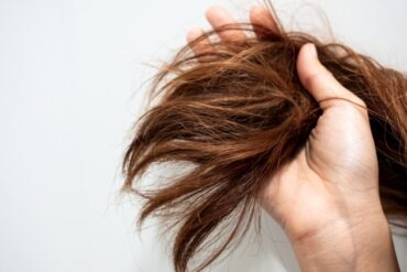 Close-up of a person's hand holding the ends of long, dry, reddish-brown hair in front of a light gray background.