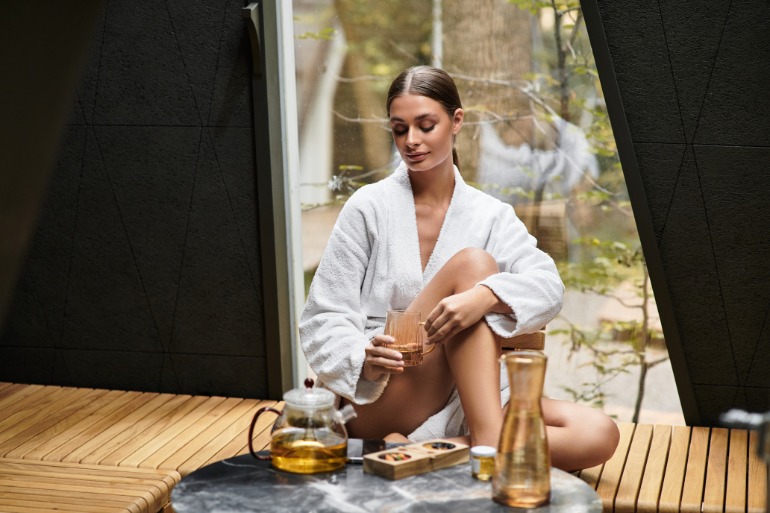Spa guest in a white robe sits by a window holding a glass of tea beside a table with a teapot and carafe.