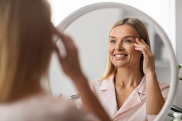A smiling woman inspecting herself with a hand below one eye while facing a mirror in a bright bathroom with white walls.