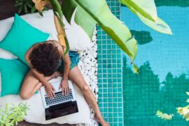 Overhead view of a person working on a laptop beside a turquoise swimming pool surrounded by tropical plants.