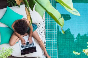 Overhead view of a person working on a laptop beside a turquoise swimming pool surrounded by tropical plants.