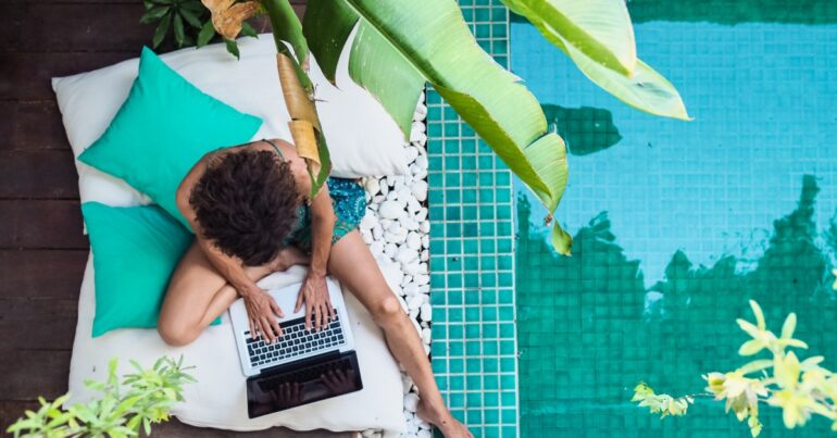 Overhead view of a person working on a laptop beside a turquoise swimming pool surrounded by tropical plants.