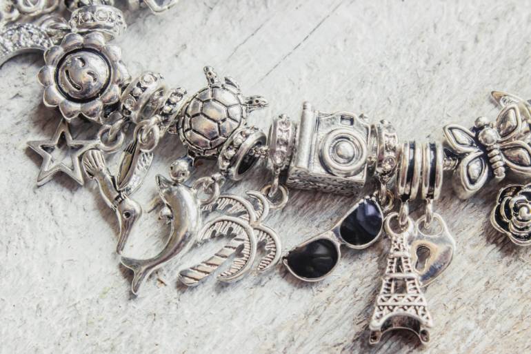 A silver charm bracelet on white wood, with pendants shaped like a turtle, sunglasses, a tower, and a butterfly.