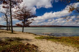 An empty picnic table sits in a sandy area next to a large lake on a sunny fall day. Leaves litter the ground.