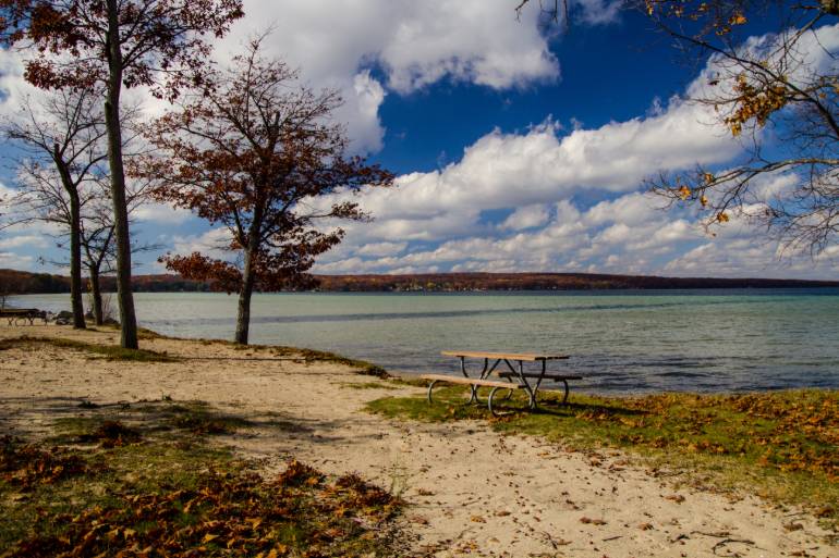 An empty picnic table sits in a sandy area next to a large lake on a sunny fall day. Leaves litter the ground.