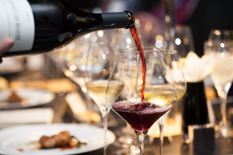 A person pours red wine from a bottle into a wine glass on a dining table set with food and additional glasses.
