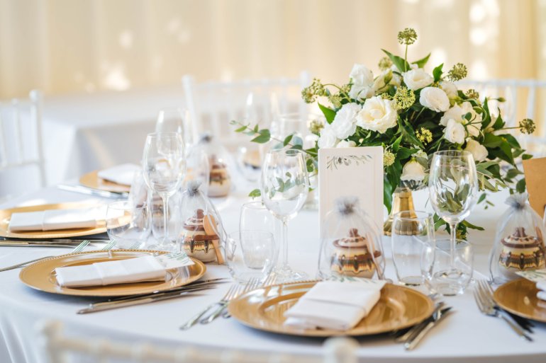 Elegant wedding table with gold place settings, white linens, glassware, and a lush centerpiece of white flowers.