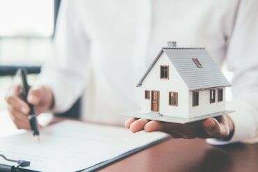 A person holds a miniature white house with a gray roof in one hand while signing paperwork with a pen.