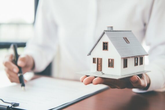 A person holds a miniature white house with a gray roof in one hand while signing paperwork with a pen.