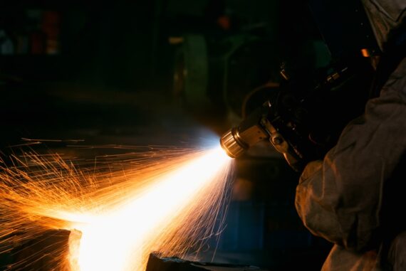 A worker uses a thermal spray gun to apply a bright coating to a metal surface, sending orange sparks across a dark workshop.