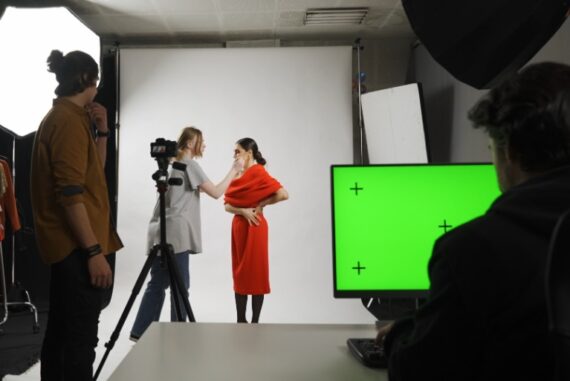 A woman in a red dress standing before a white backdrop as another woman adjusts her makeup near a bright green monitor.