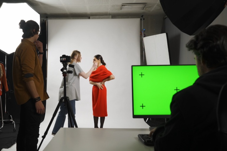 A woman in a red dress standing before a white backdrop as another woman adjusts her makeup near a bright green monitor.