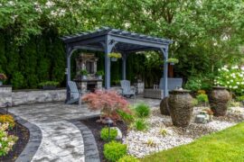 A landscaped backyard with a gray pergola, stone fireplace, patio chairs, decorative fountains, and lush greenery.