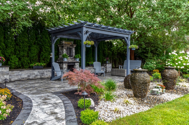 A landscaped backyard with a gray pergola, stone fireplace, patio chairs, decorative fountains, and lush greenery.