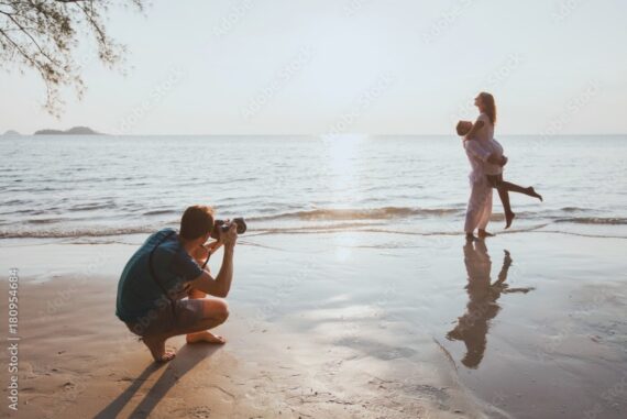 Photographer crouches on a sandy beach as two women pose together at the shoreline in warm evening light.